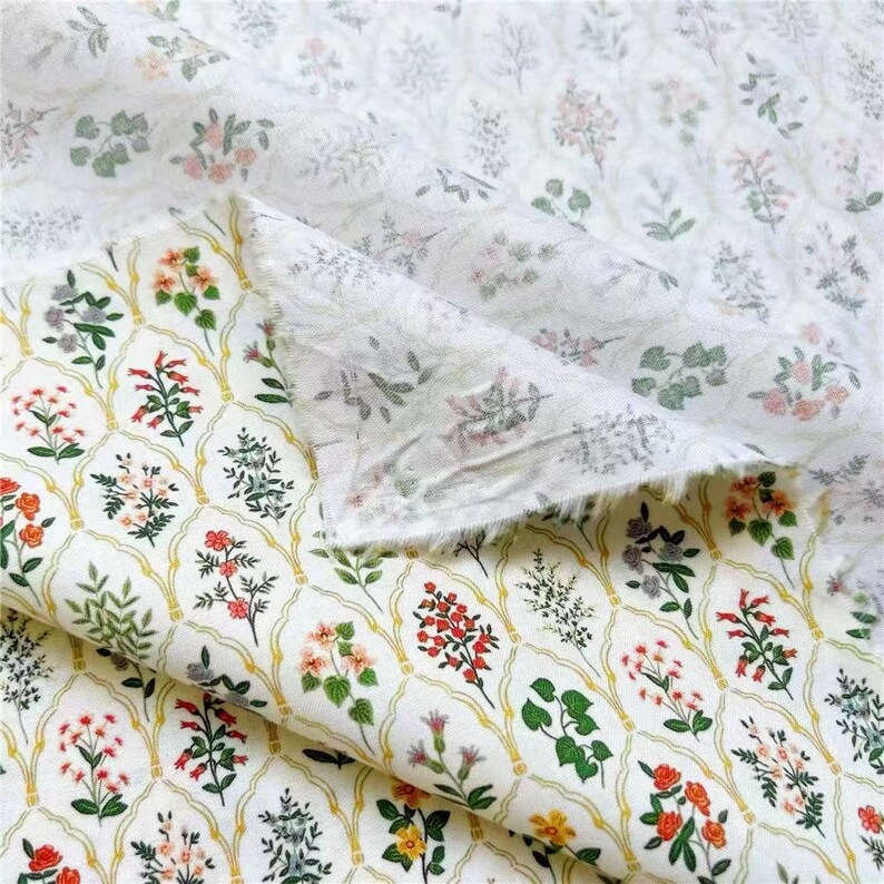 May include: White cotton fabric with a floral print. The fabric features a repeating pattern of small flowers and leaves in shades of pink, red, green, and yellow. The fabric is folded in half, revealing the back side of the fabric, which is a solid white.