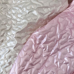 May include: Two sections of quilted fabric, one in soft white and the other in pale pink. Both fabrics feature a repeating heart pattern created by the stitching. The material has a shiny, satin-like finish, reflecting light.