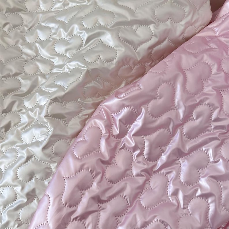 May include: Two sections of quilted fabric, one in soft white and the other in pale pink. Both fabrics feature a repeating heart pattern created by the stitching. The material has a shiny, satin-like finish, reflecting light.