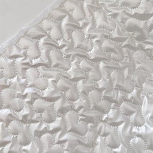 May include: Close-up of a white, quilted fabric featuring a heart pattern. The fabric has a shiny, satin-like appearance and a textured surface. The hearts are stitched with a dotted line, creating a raised effect. The edge of the fabric has a white trim.