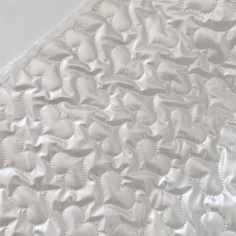 May include: Close-up of a white, quilted fabric featuring a heart pattern. The fabric has a shiny, satin-like appearance and a textured surface. The hearts are stitched with a dotted line, creating a raised effect. The edge of the fabric has a white trim.