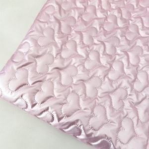May include: A close-up of a pink quilted fabric with a heart pattern. The fabric has a shiny, satin-like appearance. The hearts are outlined with a dotted stitch, creating a textured effect. The fabric is folded, showing the underside.