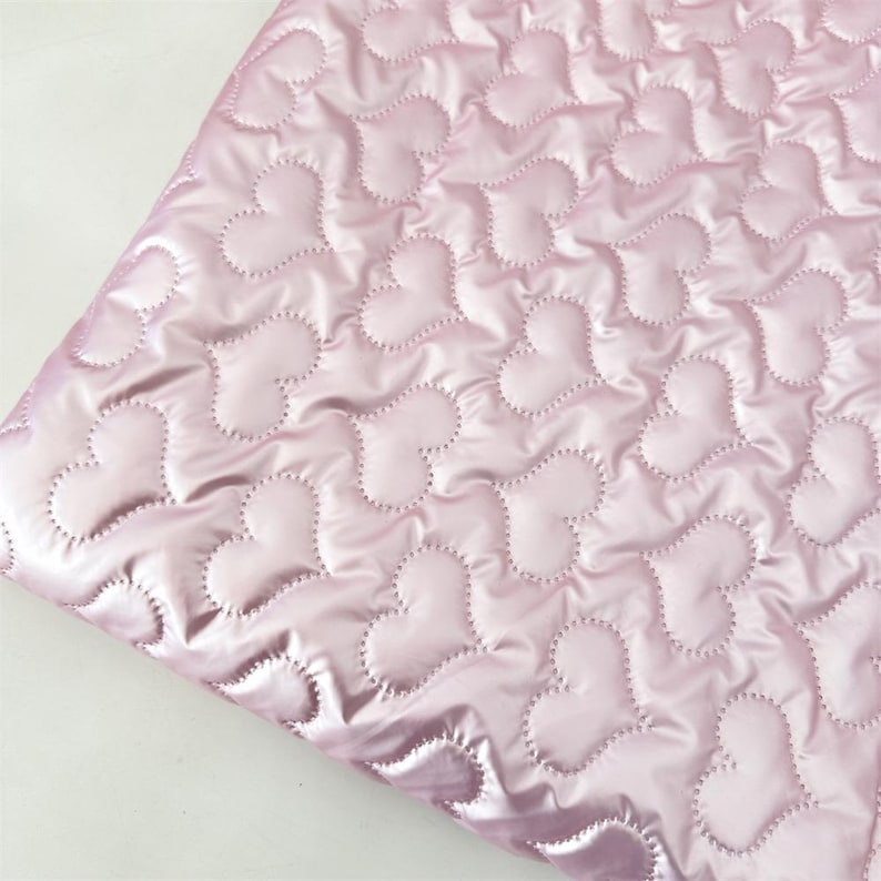 May include: A close-up of a pink quilted fabric with a heart pattern. The fabric has a shiny, satin-like appearance. The hearts are outlined with a dotted stitch, creating a textured effect. The fabric is folded, showing the underside.