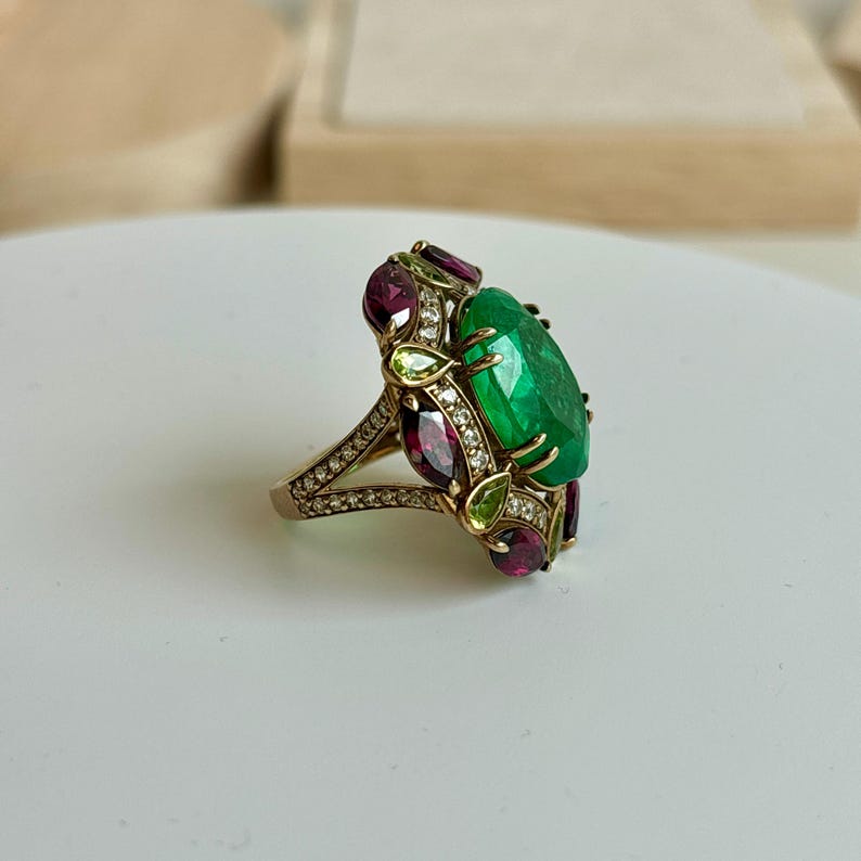 K&ouml;nnte beinhalten: Ein goldener Ring mit einem gro&szlig;en, oval geschliffenen Smaragd im Zentrum. Der Ring ist mit kleineren Edelsteinen in Rot und Gr&uuml;n verziert, die den Smaragd umgeben. Das Band ist mit kleinen, funkelnden Akzenten versehen.