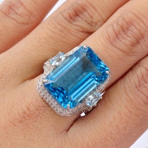 Puede incluir: Un gran anillo rectangular con una piedra preciosa azul y una banda plateada. El anillo presenta una piedra azul central de talla esmeralda, flanqueada por piedras de acento azul claro más pequeñas. La banda está detallada con pequeñas piedras transparentes.