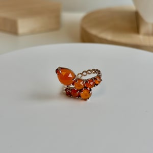 May include: A unique ring featuring a series of orange gemstones. The ring has a spiral design with a large, teardrop-shaped stone at the top and smaller, faceted stones along the band. The ring is set on a gold-colored metal band.