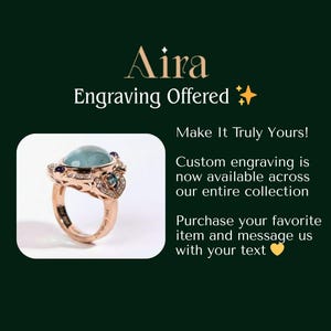K&ouml;nnte beinhalten: Ein ros&eacute;goldener Ring mit einem gro&szlig;en, hellblauen Edelstein und kleineren lila und blauen Steinen. Der Ring hat ein detailliertes Design und ist vor einem dunkelgr&uuml;nen Hintergrund platziert. Der Text auf dem Bild lautet "Aira Gravur angeboten."