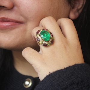 K&ouml;nnte beinhalten: Ein auff&auml;lliger Ring mit einem gro&szlig;en, ovalen Smaragd in der Mitte, umgeben von kleineren roten und klaren Edelsteinen. Der Ring hat ein goldfarbenes Band und eine Fassung. Der Ring befindet sich an einem Finger.