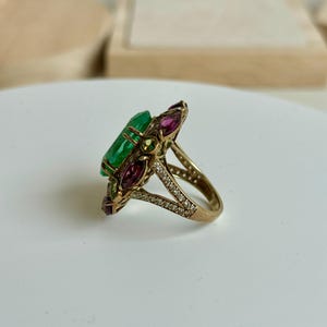 K&ouml;nnte beinhalten: Ein goldener Ring mit einem gro&szlig;en, rechteckigen Smaragd-Mittelstein, umgeben von kleineren, farbigen Edelsteinen. Das Ringband ist mit kleinen, klaren Steinen besetzt. Das Design ist aufwendig und detailliert und zeigt einen Vintage- oder Antik-Stil.