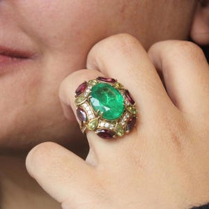 K&ouml;nnte beinhalten: Ein goldener Ring mit einem gro&szlig;en, oval geschliffenen Smaragd-Mittelstein, umgeben von kleineren roten und gr&uuml;nen Edelsteinen und Akzentdiamanten. Der Ring hat eine verzierte, detaillierte Fassung.