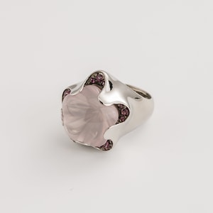May include: A silver ring featuring a large, light pink gemstone with a flower-like design. Small, dark pink gemstones are set around the central stone. The ring has a smooth, polished finish and a wide band.