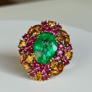 May include: A gold ring featuring a large, oval-cut emerald center stone, surrounded by a cluster of smaller, round rubies, and teardrop-shaped citrines. The gemstones are set in a gold band, creating a vibrant and luxurious piece of jewelry.