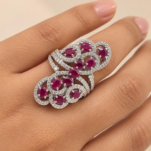 May include: A silver-toned statement ring featuring an elongated, swirling filigree design encrusted with numerous small, clear pavé stones and accented with multiple oval-cut, deep pinkish-red rubies arranged in a cluster pattern.