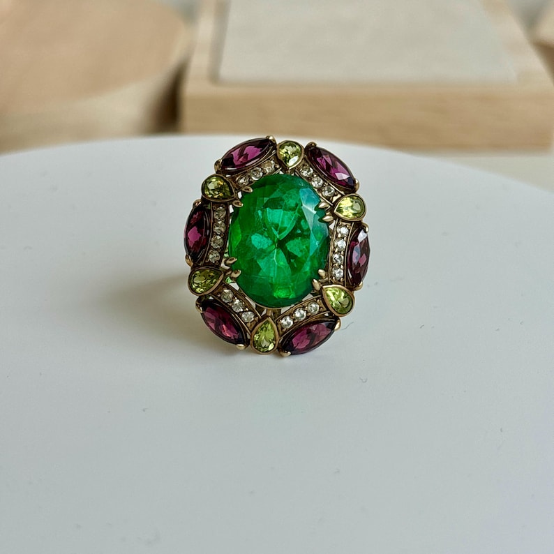 K&ouml;nnte beinhalten: Ein goldfarbener Ring mit einem gro&szlig;en, oval geschliffenen, leuchtend gr&uuml;nen Edelstein in der Mitte. Der Edelstein ist von kleineren, runden, klaren Steinen und abwechselnd oval geschliffenen, tiefroten und hellgr&uuml;nen Edelsteinen umgeben.