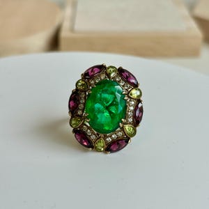 K&ouml;nnte beinhalten: Ein goldener Ring mit einem gro&szlig;en, oval geschliffenen Smaragd als Mittelstein. Der Smaragd ist von kleinen, runden Diamanten und abwechselnd oval geschliffenen, tiefroten und hellgr&uuml;nen Edelsteinen umgeben. Der Ring liegt auf einer wei&szlig;en Oberfl&auml;che.