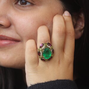 K&ouml;nnte beinhalten: Ein aufwendiger Ring mit einem zentralen gr&uuml;nen Smaragd, umgeben von kleineren roten und klaren Edelsteinen. Der Ring, gefasst in Gold, wird an einem Finger getragen.