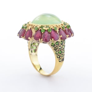 May include: A gold ring featuring a large, light green, oval-shaped center stone. The ring is adorned with a halo of red teardrop-shaped stones and smaller green stones. The band is also embellished with green stones.