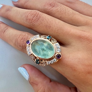 Puede incluir: Un anillo de oro con una gran piedra preciosa ovalada verde rodeada de piedras preciosas más pequeñas transparentes y azules. El anillo está engastado con un diseño decorativo y tiene una piedra preciosa púrpura a cada lado de la piedra principal.