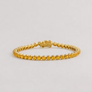 May include: A gold-toned tennis bracelet featuring a continuous row of round, faceted yellow stones set in a metallic frame. The bracelet includes a secure box clasp mechanism at the center, showcasing a uniform design of sparkling amber-colored gems.