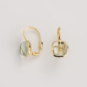 May include: A pair of gold-colored leverback earrings. Each earring features a light green, oval-shaped gemstone. The gemstones are set in a gold bezel setting. The earrings are displayed against a white background.