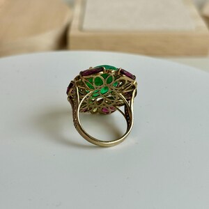 K&ouml;nnte beinhalten: Ein goldener Ring mit einem gro&szlig;en, ovalen gr&uuml;nen Edelstein in der Mitte, umgeben von kleineren, rechteckigen roten und rosa Edelsteinen. Der Ring hat ein aufwendiges, offenes Design und eine polierte Oberfl&auml;che.