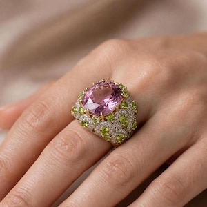 May include: A gold ring featuring a large oval-cut pink centre stone surrounded by a cluster of smaller round green stones and tiny clear pavé diamonds set in a decorative metal frame that covers the top portion of the finger.