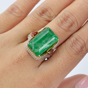 Puede incluir: Anillo con una gran gema verde rectangular, flanqueada por dos gemas rojas más pequeñas. El anillo tiene una banda de oro y plata con un diseño intrincado y detallado, destacando la esmeralda.