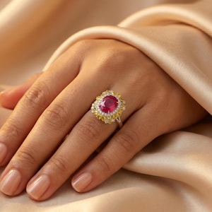 Könnte beinhalten: Ein silberner Ring mit einem großen, ovalen Rubin. Der Rubin ist von kleinen, gelben Edelsteinen und einem Rand aus weißen Edelsteinen umgeben. Der Ring befindet sich vor einem weichen, goldenen Hintergrund.