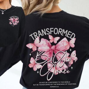 May include: Black long-sleeve t-shirt with a pink bow and butterflies graphic. The text "TRANSFORMED By God's Grace" is printed on the shirt. The scripture "Romans 12:2" is also printed on the shirt.