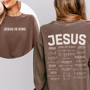 Op de afbeelding: Een espresso-kleurig sweatshirt met lange mouwen en de tekst JESUS IS KING op de voorkant. De achterkant toont het woord JESUS in grote letters boven een lijst met diverse religieuze namen en titels in verschillende witte lettertypes.