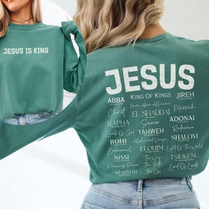 May include: A long-sleeve light green sweatshirt featuring the text "JESUS IS KING" on the front. The back displays the word "JESUS" in large white letters above a list of various names and titles for Jesus printed in multiple white fonts.
