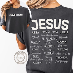 Op de afbeelding: Een antracietgrijs T-shirt met korte mouwen en witte tekst. De voorkant toont "JESUS IS KING". De achterkant bevat het woord "JESUS" in grote, verweerde letters boven een lijst met diverse religieuze titels en namen in verschillende lettertypes.