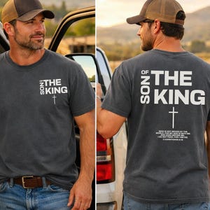 May include: A dark gray t-shirt with the text "SON OF THE KING" and a cross graphic. The shirt is worn with a brown and tan baseball cap and blue jeans. The text "WHO IS NOT MOVED BY THE WORLD, FOR MY GOD IS WITH ME I DO NOT FEAR, FOR HE IS" is also printed on the back.
