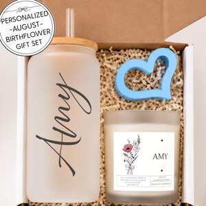 Op de afbeelding: Een geschenkset in een doos met een matglazen beker met bamboe deksel, een doorzichtig rietje en de naam Amy in zwarte letters. Een kaars in een glazen pot met een bloemenillustratie, de naam Amy en tekst. Een lichtblauw object in de vorm van een hart.