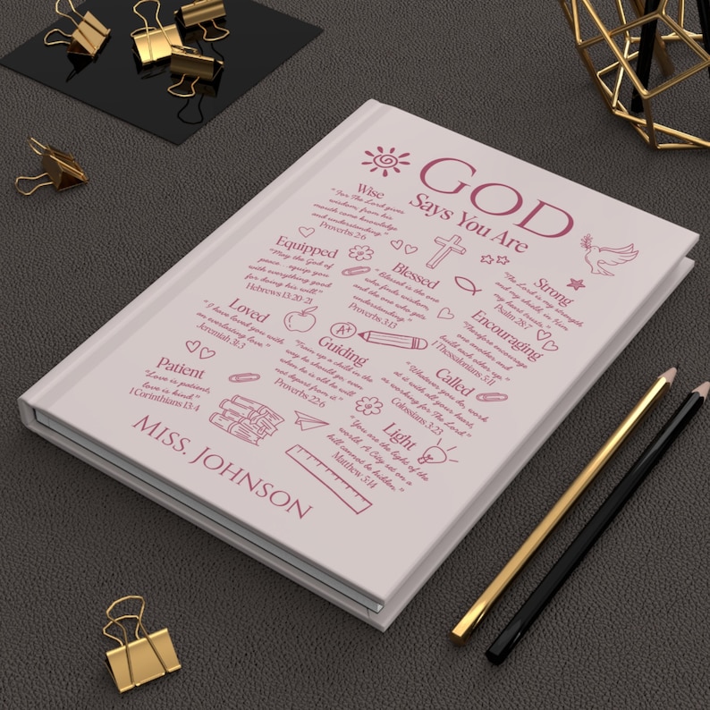 May include: A light pink journal with the words "GOD Says You Are" and inspirational phrases in a handwritten style. The journal has a white border and is accompanied by gold and black pencils, and gold binder clips.