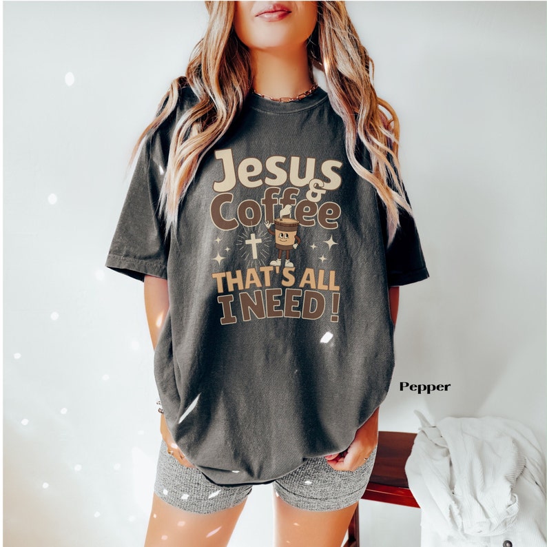 May include: A dark gray t-shirt with the text "Jesus & Coffee That's All I Need!" in beige and brown lettering. A cartoon coffee cup with a cross is featured in the design.