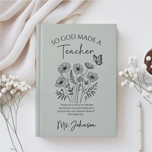 May include: A light gray journal with the text "SO GOD MADE A Teacher" and a floral and butterfly design. The journal also includes a quote from Proverbs 22:6 and the name "Ms. Johnson". A pen and a cup of coffee are visible.