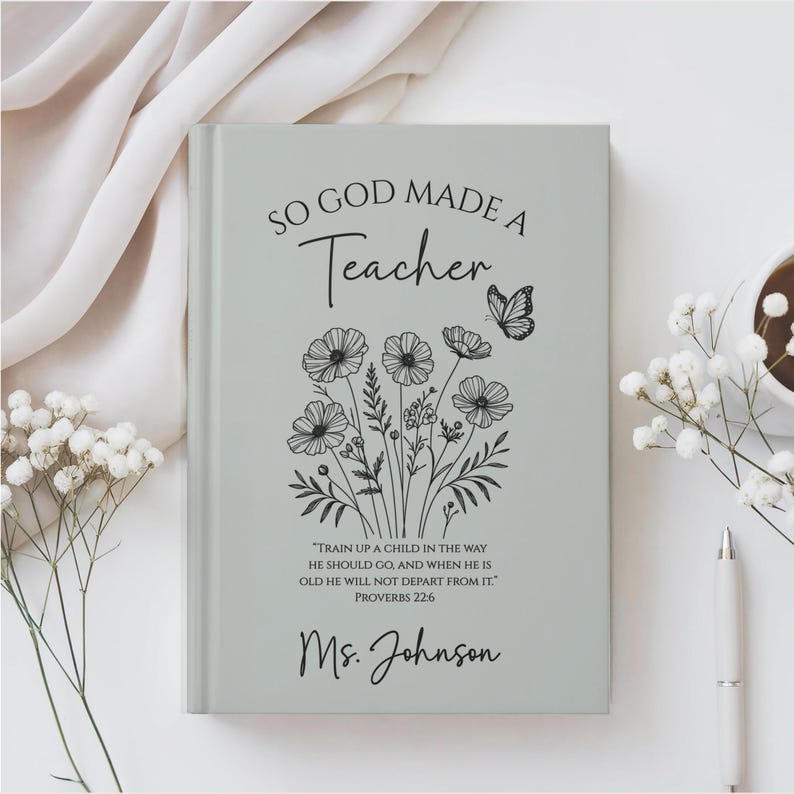 May include: A light gray journal with the text "SO GOD MADE A Teacher" and a floral and butterfly design. The journal also includes a quote from Proverbs 22:6 and the name "Ms. Johnson". A pen and a cup of coffee are visible.