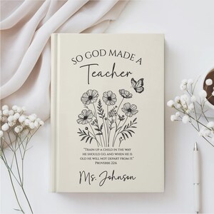 May include: A cream-colored journal with the text "SO GOD MADE A Teacher" and a floral illustration. Below the text is a quote from Proverbs 22:6 and the name "Ms. Johnson". A butterfly is also featured in the design.
