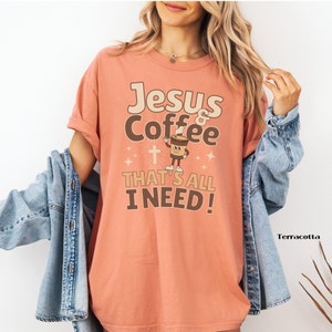 May include: A terracotta colored t-shirt with the text "Jesus & Coffee That's All I Need!" in brown and white lettering. A cartoon coffee cup with a cross is in the center of the design.