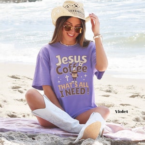 May include: A woman wearing a purple t-shirt with the text "Jesus & Coffee That's All I Need!" and a cartoon image of a coffee cup with a cross. She is wearing a straw cowboy hat and white boots. The background is a beach.