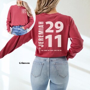 Op de afbeelding: Crimson langarm T-shirt met de tekst "JEREMIAH 29:11 FOR I KNOW THE PLANS I HAVE FOR YOU" gedrukt op de achterkant in wit. Het shirt wordt gedragen door iemand die een blauwe jeans draagt.