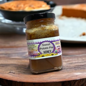 May include: A glass jar of honey with a purple and white label that reads "Sweet Potato Pie Honey". The jar is sitting on a wooden surface.