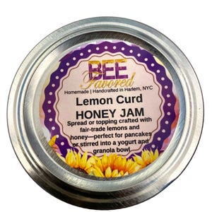 May include: A jar of Bee Favored Lemon Curd Honey Jam. The label features text and a purple and white design with sunflowers. The jar is made of metal and contains a spread crafted with fair-trade lemons and honey, perfect for pancakes or yogurt.