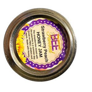 May include: A jar lid with a label that reads "Strawberry Peach Honey Jam." The label has a purple and gold design with text that says "BEE Favored." The lid is silver and the label includes text about the ingredients.