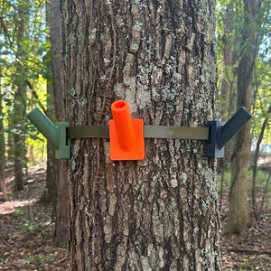 May include: A tree-mounted gear holder system featuring an olive green, a bright orange, and a dark gray angled tube attachment secured to a tree trunk by a flat, adjustable olive green nylon strap that wraps around the bark.