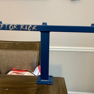 May include: A blue metal rack with the text "RAPTOR RACK" in silver lettering. The rack has a T-shape design with a horizontal bar and a vertical support. The base has four holes for mounting.
