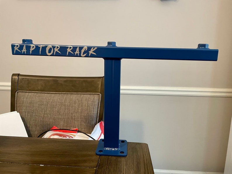 May include: A blue metal rack with the text "RAPTOR RACK" in silver lettering. The rack has a T-shape design with a horizontal bar and a vertical support. The base has four holes for mounting.