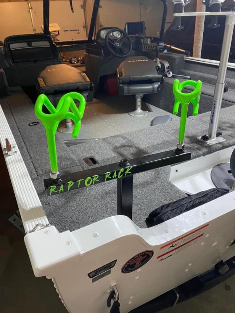 May include: A boat interior with a gray carpeted floor and black seats. The boat features a black metal Raptor Rack with two neon green rod holders. The text "RAPTOR RACK" is visible on the rack.