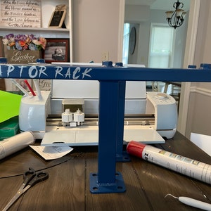 May include: A blue metal rack with the text "RAPTOR RACK" in white. The rack has a T-shaped design with a horizontal bar and a vertical support. The rack is on a wooden table, with a white cutting machine and craft supplies in the background.